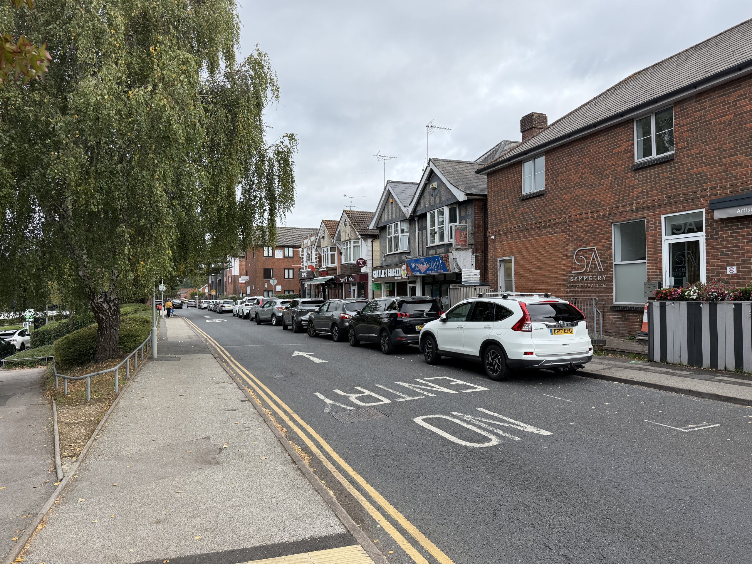 Tree-lined shopping street in Broadstone, Poole, Dorset, with local businesses, parked cars and pedestrian pavements.