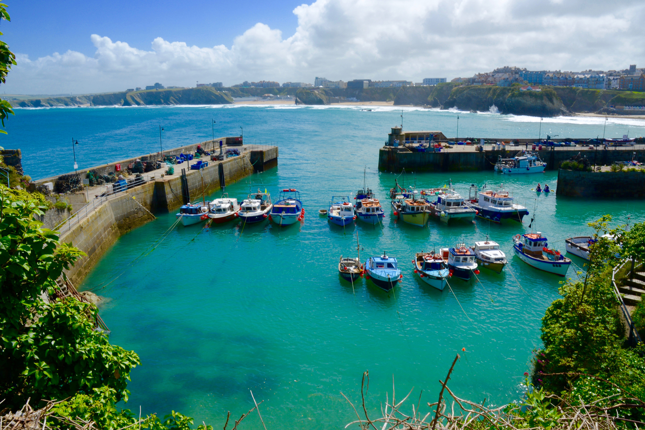 Newquay Harbour in Cornwall with colourful fishing boats moored in turquoise waters and the town's coastline in the background.