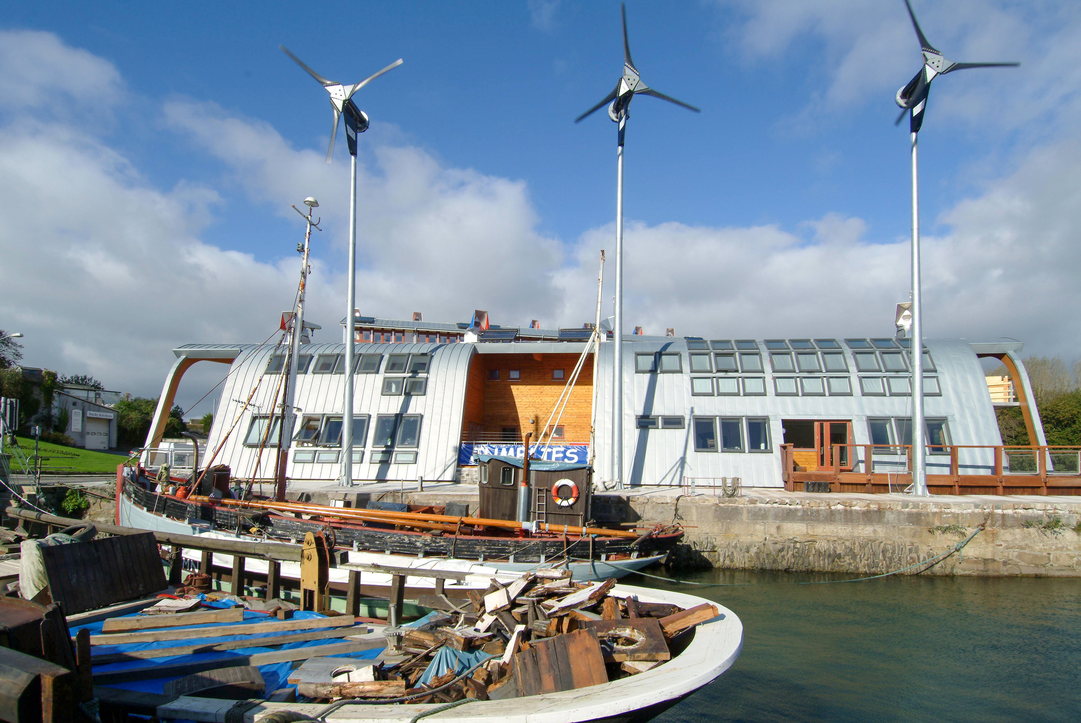 Jubilee Wharf waterfront building in Penryn, Cornwall, with wind turbines and traditional boats moored alongside.