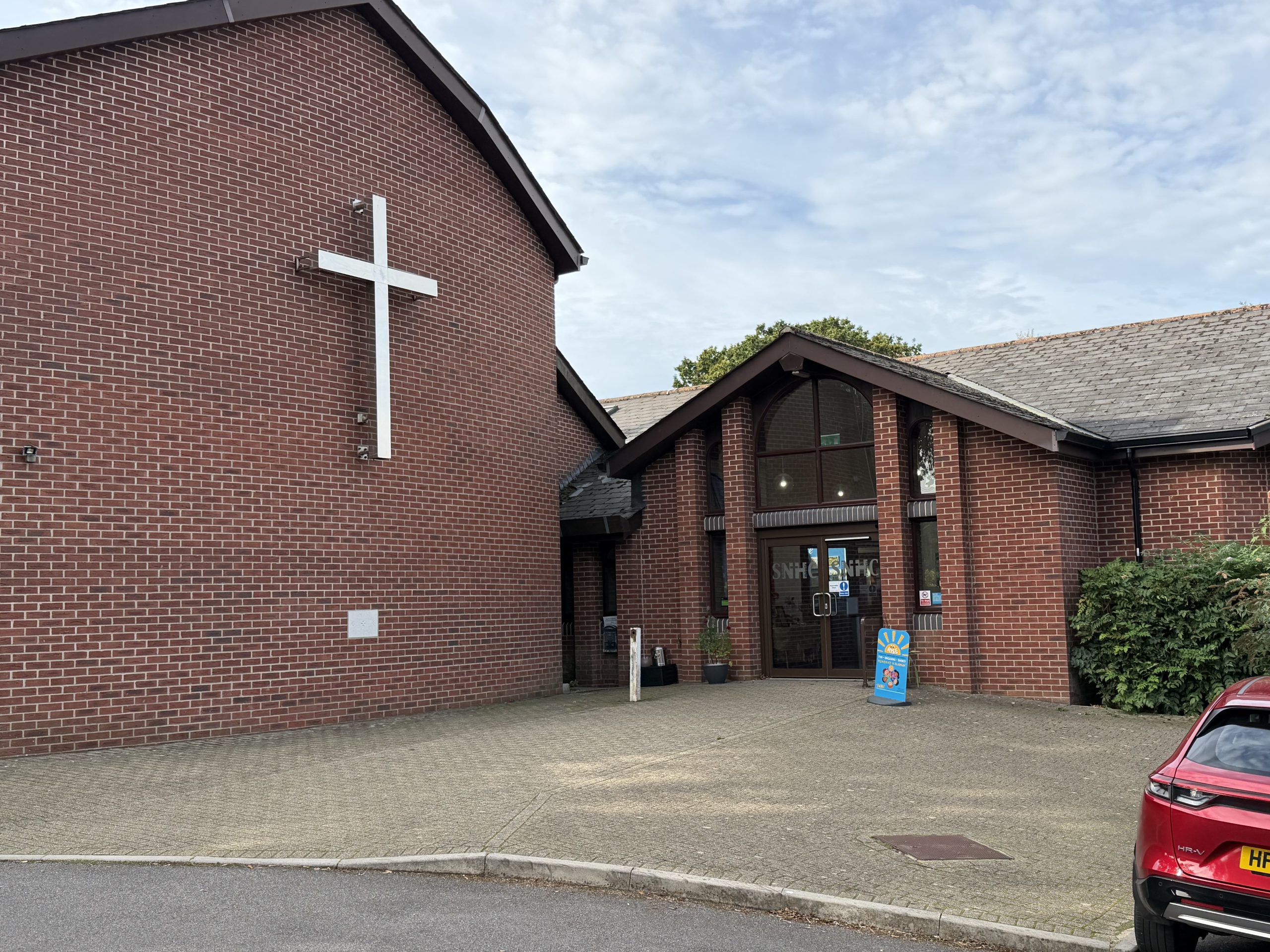 Exterior of St Nicholas Church in Corfe Mullen, Dorset, with its red-brick walls and prominent white cross.
