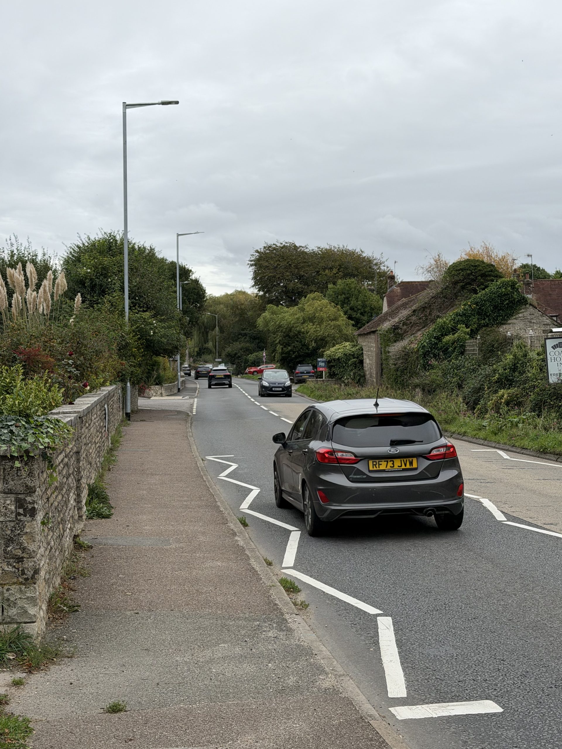 Village road through Winterbourne Abbas, Dorset, with traditional stone buildings, houses and greenery.